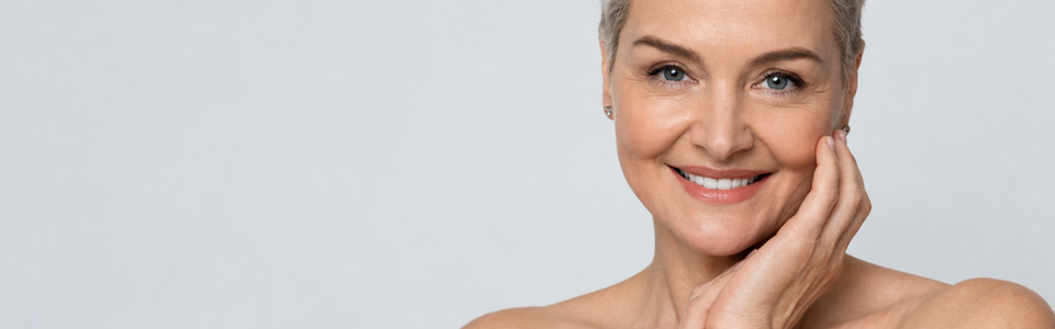 Moden kvinde med naturlig hud og et roligt smil mod en lys, ensfarvet baggrund – illustrerer behandlinger af rosacea, pigmentforandringer, karsprængninger og ar.